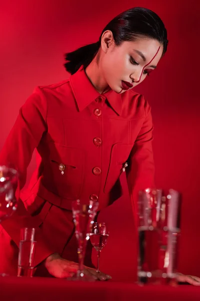 Morena mujer asiática en chaqueta de moda y maquillaje creativo de pie cerca de gafas borrosas sobre fondo rojo - foto de stock