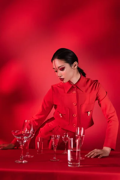 Брюнетка азиатская женщина в модном наряде и творческом облике рядом с различными очками с водой на красном абстрактном фоне — стоковое фото