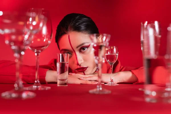 Jovem mulher asiática com maquiagem criativa no rosto dividido com linha perto de copos borrados de água no fundo vermelho — Fotografia de Stock