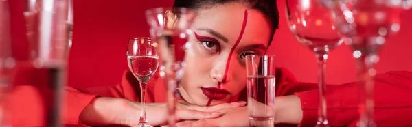 Mujer joven con maquillaje artístico mirando a la cámara cerca de vasos borrosos de agua aislada en rojo, pancarta - foto de stock