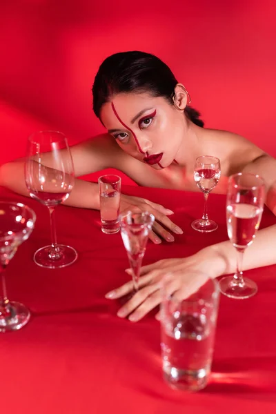 Mujer asiática desnuda con maquillaje creativo posando cerca de diferentes gafas con agua sobre fondo rojo - foto de stock
