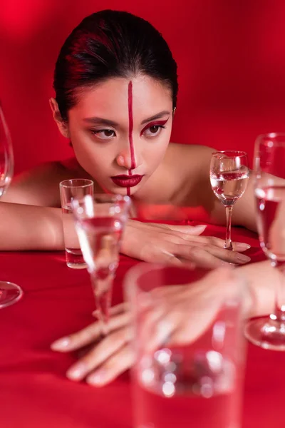 Sensual mulher asiática com visagem criativa e ombros nus posando perto de óculos de água no fundo vermelho — Fotografia de Stock