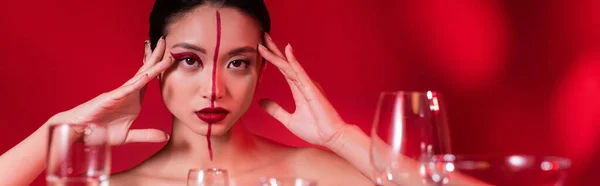 Young asian woman with naked shoulders and creative makeup touching face on red background, banner — Stock Photo