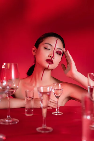 Brunette asian woman with closed eyes and artistic visage near blurred glasses on red background — Stock Photo