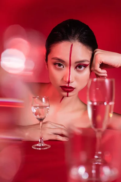 Retrato de mulher asiática com maquiagem no rosto dividido com linha olhando para a câmera perto de óculos borrados no fundo vermelho — Fotografia de Stock