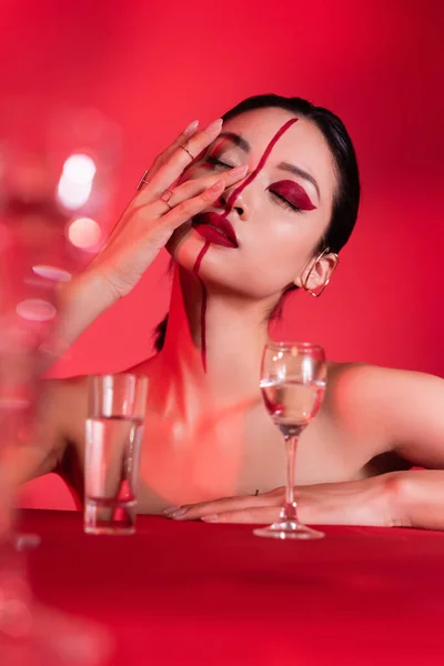 Femme asiatique avec les yeux fermés toucher le visage avec maquillage créatif près de lunettes floues sur fond rouge — Photo de stock