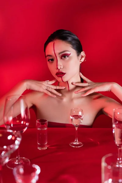 Elegante mulher asiática com ombros nus e visagem artística tocando pescoço perto de vários óculos com água no fundo vermelho — Fotografia de Stock