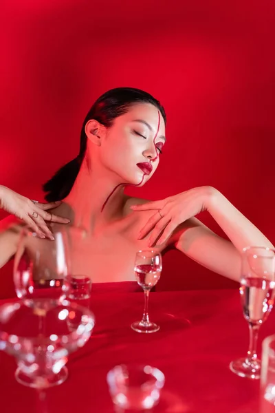 Sensual mulher asiática com olhos fechados e visagem criativa tocando ombros nus perto de óculos borrados no fundo vermelho — Fotografia de Stock