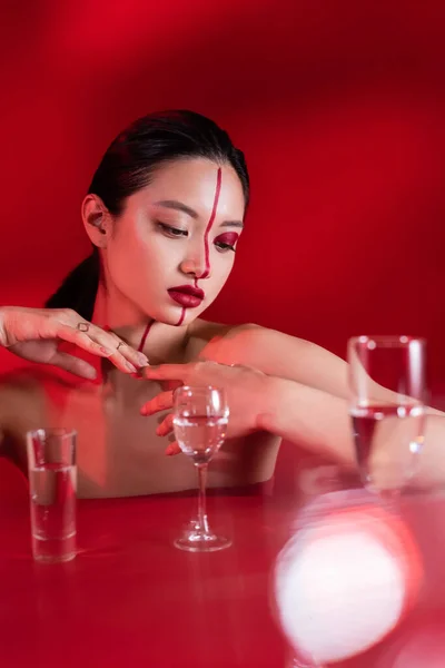 Junges asiatisches Model mit nackten Schultern und kreativem Gesicht posiert in der Nähe einer verschwommenen Brille auf rotem Hintergrund — Stockfoto