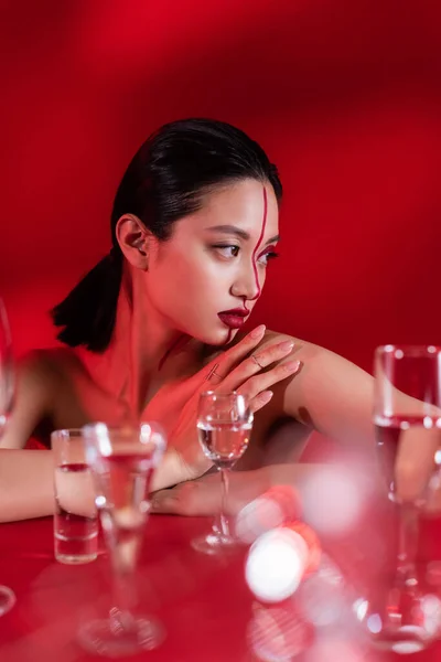 Desnudo asiático mujer con maquillaje y línea en cara mirando lejos cerca borrosa gafas en rojo fondo - foto de stock