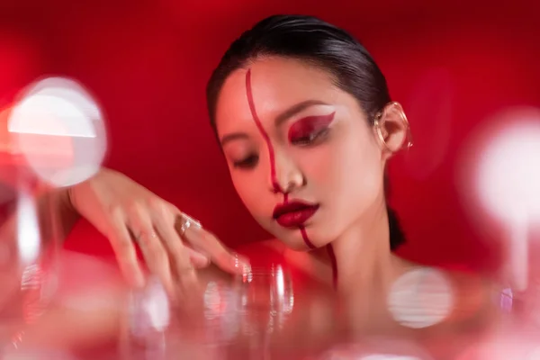 Morena mujer asiática con rostro artístico cerca de gafas borrosas sobre fondo rojo — Stock Photo