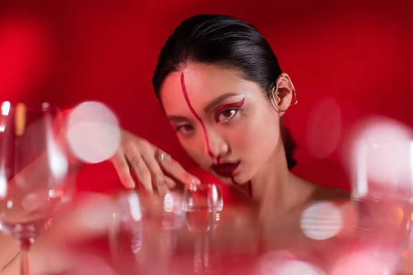 Jovem mulher asiática com maquiagem criativa olhando para a câmera perto de óculos borrados no fundo vermelho — Fotografia de Stock