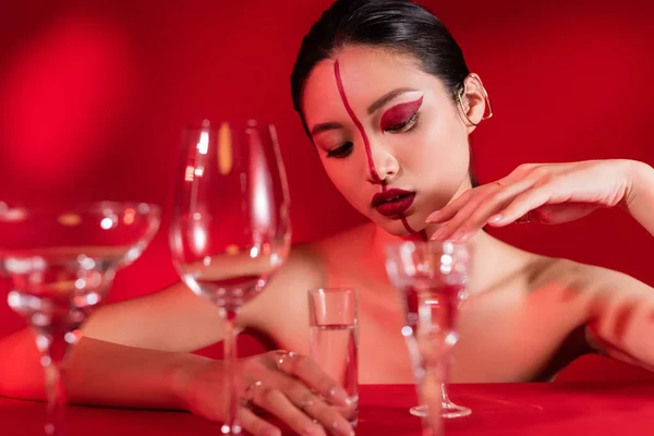 Seductive asian woman with naked shoulders and artistic visage touching glass of water on red background — Stock Photo