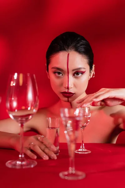 Nua mulher asiática com maquiagem criativa e linha no rosto tocando vidro com água pura no fundo vermelho — Fotografia de Stock