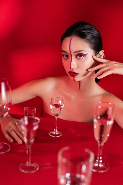 Mulher asiática com visagem criativa brilhante e mão perto do rosto olhando para a câmera perto de vários óculos no fundo vermelho — Fotografia de Stock