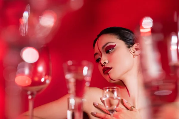 Low angle view of nude asian woman with bright creative visage touching bare shoulder near blurred glasses on red background — Stock Photo