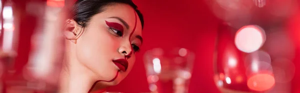 Portrait of asian woman with creative makeup on face divided with line near blurred glasses on red background, banner — Stock Photo