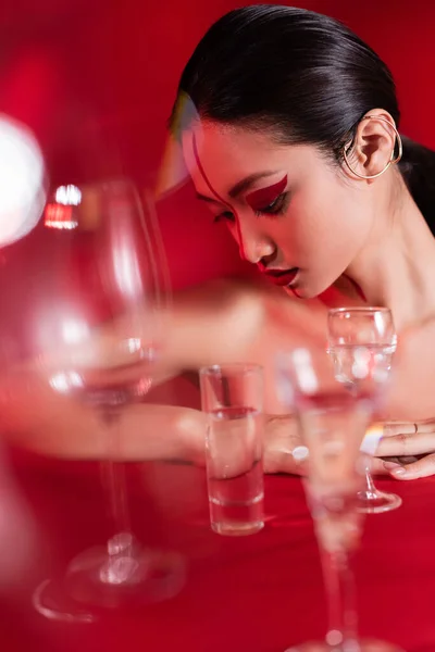 Nua mulher asiática com manguito de orelha e visagem criativa perto de copos borrados de água no fundo vermelho — Fotografia de Stock