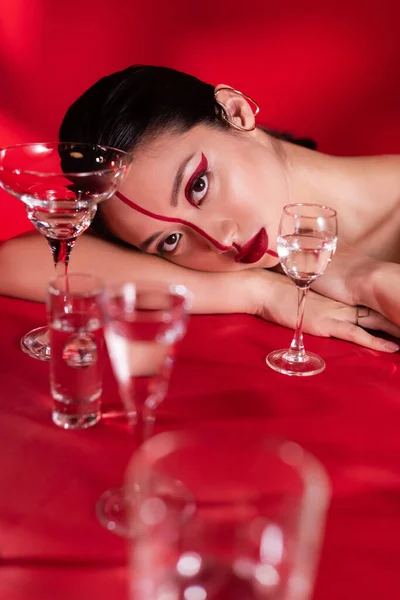 Чувственная азиатская женщина с художественным макияжем лежит рядом с различными очками с водой на красном фоне — стоковое фото