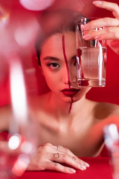 Nua mulher asiática segurando vidro de água pura perto do rosto com visagem criativa em primeiro plano borrado — Fotografia de Stock