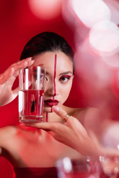 Mujer asiática con hombros desnudos y rostro creativo sosteniendo vaso de agua pura sobre fondo rojo - foto de stock