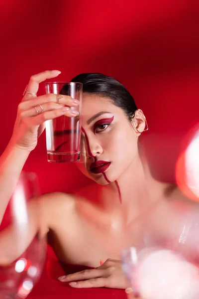 Brunette woman with makeup and bare shoulders holding glass of water near face on red background — Stock Photo