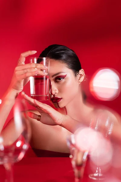 Brunette asian woman with bare shoulders and creative makeup holding glass of water near face on red background — Stock Photo