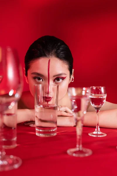 Sinnliche asiatische Frau mit artistischem Make-up blickt in die Kamera nahe Gläser mit reinem Wasser auf rotem Hintergrund — Stockfoto