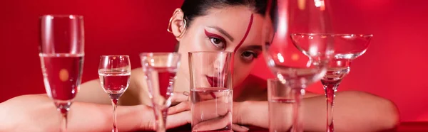 Asian woman with artistic visage and ear cuff looking at camera near glasses with pure water isolated on red, banner — Stock Photo