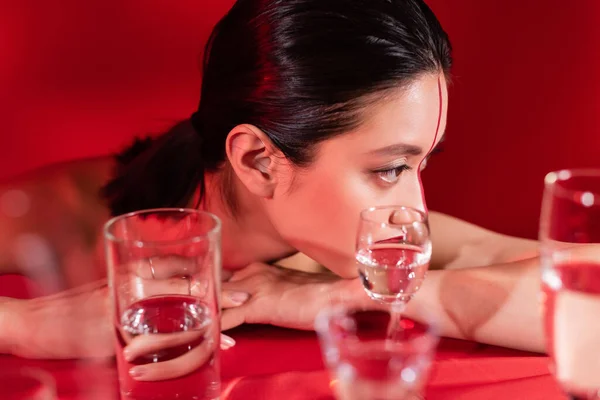 Morena asiática mulher com maquiagem olhando para longe perto de óculos borrados de água pura no fundo vermelho — Fotografia de Stock