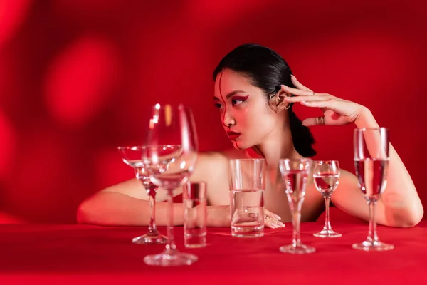 Обнаженная азиатка с творческим макияжем, смотрящая в сторону очков с прозрачной водой на красном фоне с тенью — стоковое фото