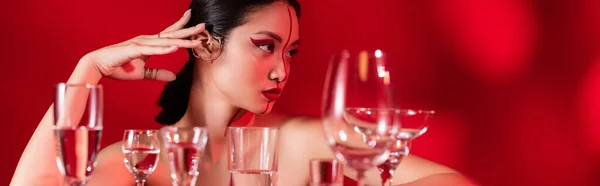 Brunette asian woman with bare shoulders and creative visage looking away near different glasses on red background, banner — Stock Photo