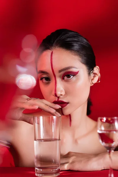 Portrait of asian woman with artistic makeup on face divided with line touching glass with pure water on red background — Stock Photo