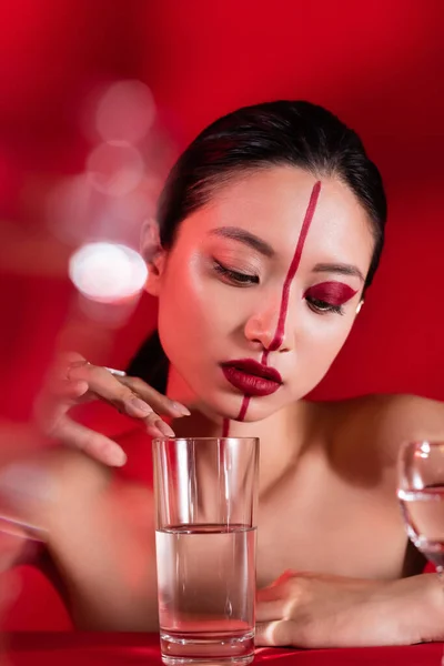 Jovem mulher asiática com maquiagem criativa e ombros nus tocando vidro com água pura no fundo vermelho — Fotografia de Stock