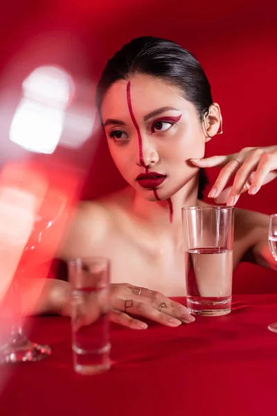 Обнаженная азиатка с художественным макияжем и ушными манжетами, смотрящая на размытые очки на красном фоне — стоковое фото