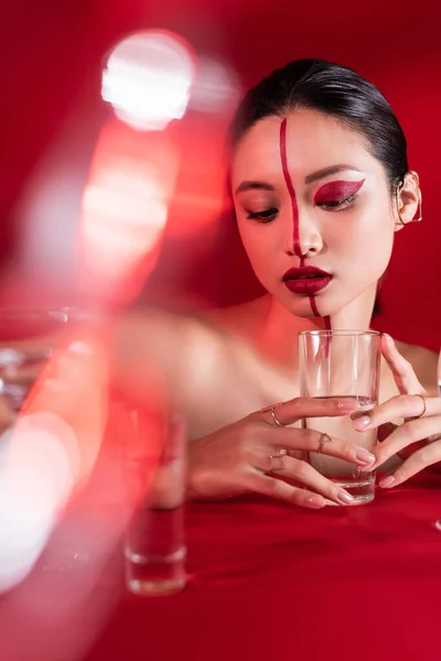 Nua mulher asiática com maquiagem artística vermelha segurando vidro de água clara em primeiro plano borrado — Fotografia de Stock