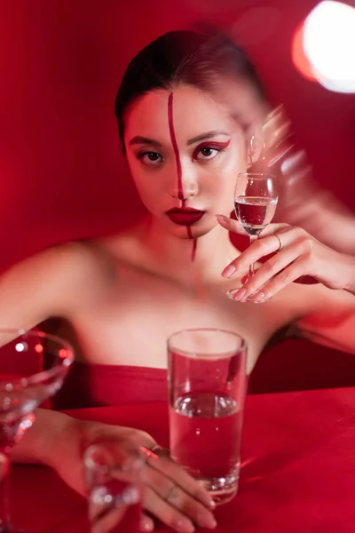 Брюнетка азиатская женщина с красным творческим обликом держа стакан чистой воды на размытом переднем плане — стоковое фото