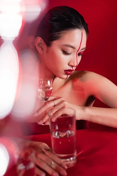 Sinnliche asiatische Frau mit kreativem Make-up, die ein Glas Wasser nahe der roten Oberfläche auf verschwommenem Vordergrund hält — Stockfoto
