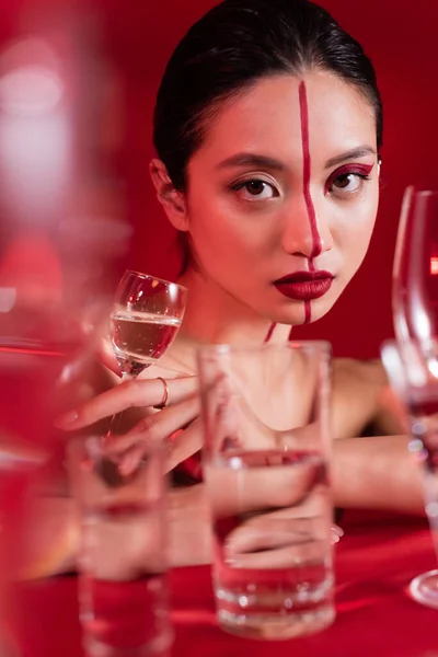 Retrato de encantadora mujer asiática con rostro creativo mirando a la cámara cerca de gafas borrosas sobre fondo rojo - foto de stock