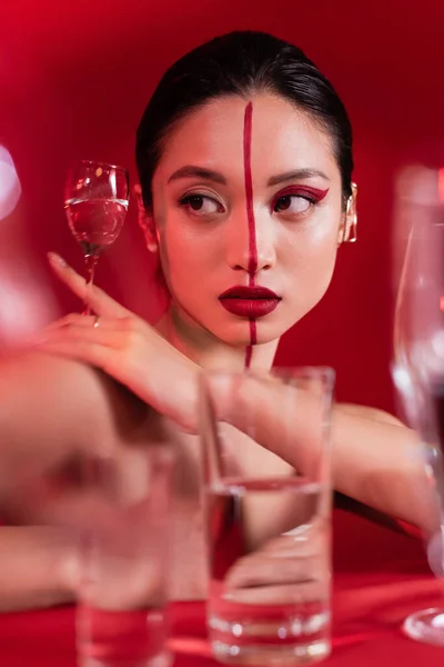 Портрет молодой азиатки с красным художественным макияжем, держащей стекло с прозрачной водой на размытом переднем плане — стоковое фото