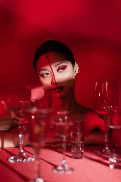 Соблазнительная азиатка с наручниками для ушей и креативным макияжем, смотрящая в свет вблизи размытых очков на красном фоне — стоковое фото
