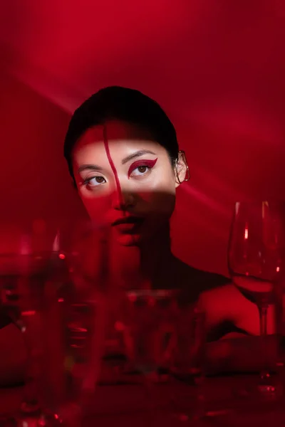 Jovem mulher asiática com maquiagem artística olhando para longe na luz perto de óculos borrados no fundo vermelho escuro — Fotografia de Stock
