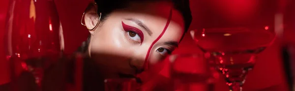 Charmante asiatische Frau mit hellem künstlerischem Make-up, die im Licht in der Nähe einer verschwommenen Brille auf rotem Hintergrund wegschaut, Banner — Stockfoto