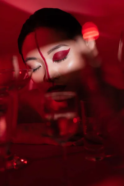 Jovem mulher asiática com olhos fechados e visagem artística na luz perto de óculos borrados no fundo vermelho escuro — Fotografia de Stock