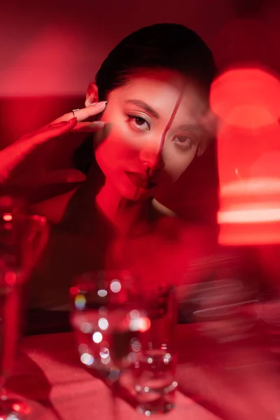 Verschwommene asiatische Frau mit kreativem Gesicht, die die Hand in der Nähe des Gesichts hält und auf rotem Hintergrund in die Kamera schaut — Stockfoto