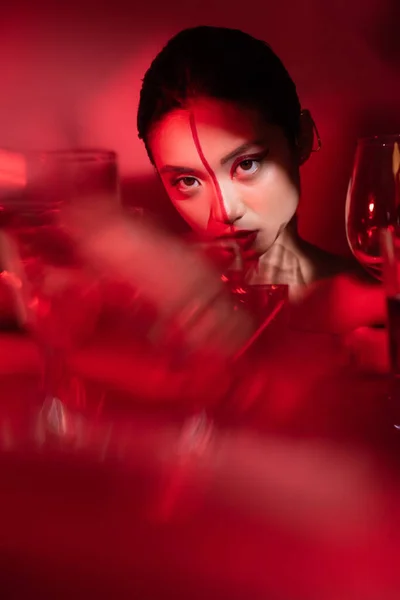 Retrato de morena mujer asiática con rostro creativo cerca de gafas borrosas sobre fondo rojo - foto de stock