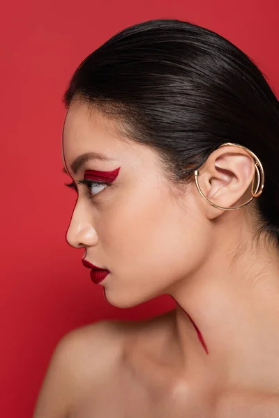 Profilo di bruna donna asiatica con visage creativo e bracciale auricolare isolato sul rosso — Foto stock