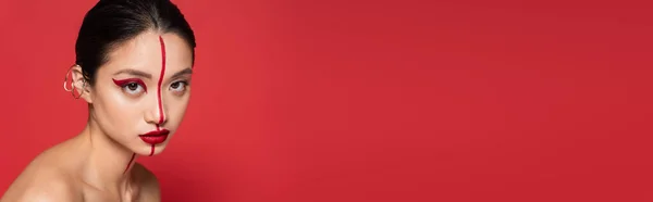 Giovane donna asiatica con visage creativo e bracciale auricolare guardando la fotocamera isolata sul rosso, banner — Foto stock