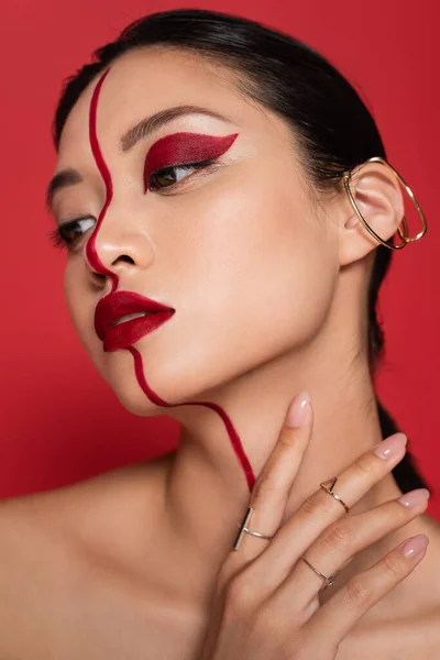 Portrait de femme asiatique brune avec maquillage créatif et boucle d'oreille manchette tenant la main près du cou isolé sur rouge — Photo de stock