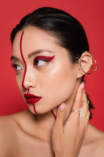 Retrato de asiático mulher com criativo visagem e orelha manguito tocando pescoço e olhando para longe isolado no vermelho — Fotografia de Stock